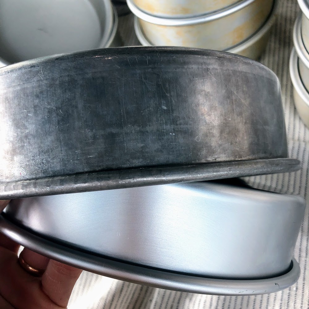 Two aluminum cake pans. One is discolored from being washed in the dishwasher. The other pan is a shiny, brand new pan.