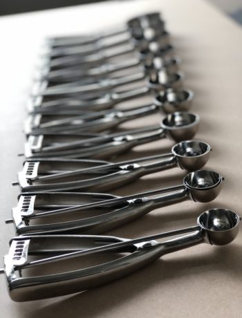 12 sizes of food dishers lined up on a brown background.