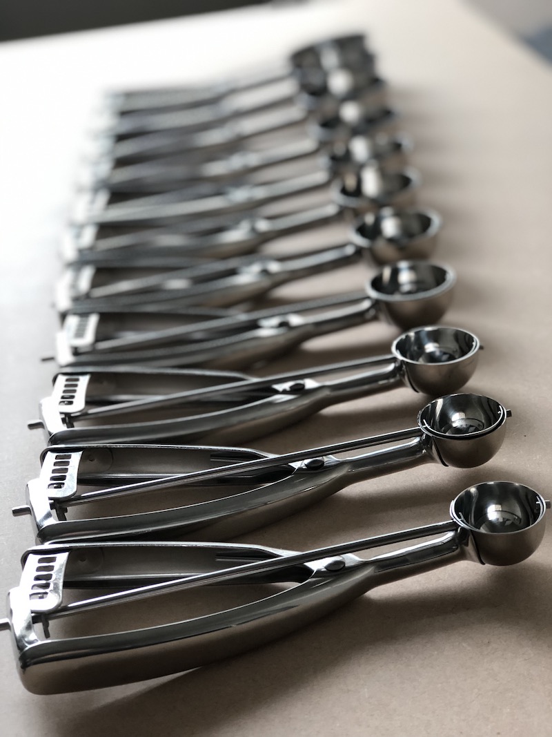 12 sizes of food dishers lined up on a brown background.