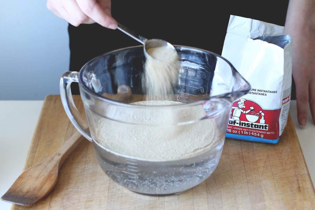 Large liquid measuring cup with sugar and yeast being sprinkled into the warm water, preparing for proofing of the yeast. 