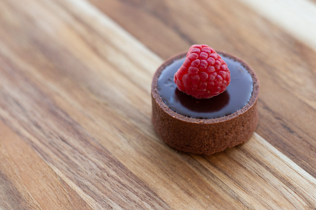 A miniature chocolate tart shell is filled with dark chocolate ganache and topped with a raspberry. 